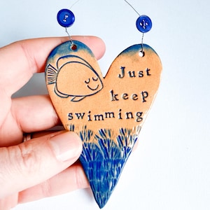 Può includere: Decorazione da parete in ceramica a forma di cuore con un'illustrazione di pesce e la scritta "Just keep swimming". Il cuore è beige con dettagli blu e un pesce blu. Due bottoni blu sono attaccati in alto per appenderlo.