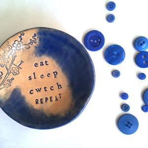 Peut inclure: Un plat en céramique bleu et beige avec un motif floral blanc et le texte "eat sleep cwtch repeat". Le plat est entouré de boutons bleus.