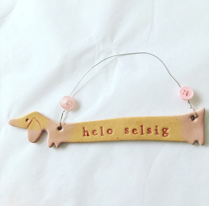 Op de afbeelding: Een keramische dachshund-vormige ornament met een roze en gele glazuur. De ornament heeft de woorden "hello selsig" erop geschreven in rood.
