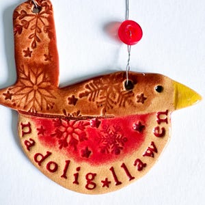 May include: A ceramic bird ornament with a red breast and yellow beak. The ornament has a brown body with snowflake and star designs. The text "nadolig llawen" is written on the bottom. A red button is attached to the top with wire.