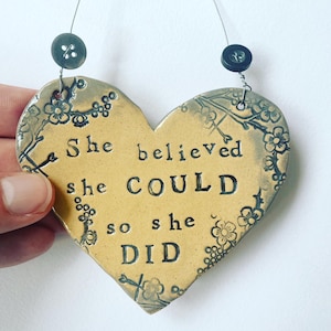 Può includere: Un ornamento in ceramica a forma di cuore giallo con un motivo floreale nero e il testo "She believed she COULD so she DID".