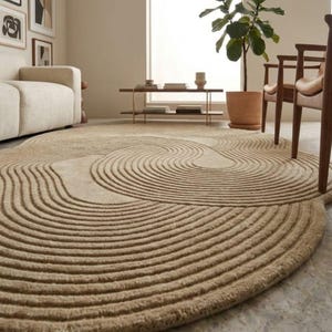 Op de afbeelding: Een beige, ovaalvormig tapijt met een getextureerd, concentrisch golfpatroon. Het tapijt ligt op een marmeren vloer in een woonkamer. Het ontwerp en de kleurstelling van het tapijt passen bij de neutrale tinten van de kamer.