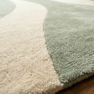 May include: Close-up of a soft, textured rug with a flowing design. The rug features a cream-colored central wave flanked by sections of sage green. The plush fibers create a cozy, inviting feel, suitable for home decor.