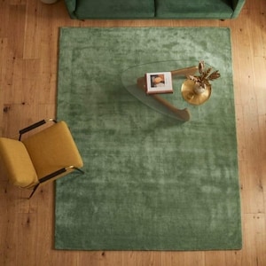 Könnte beinhalten: Draufsicht auf ein Wohnzimmer mit einem großen, rechteckigen, grünen Teppich. Ein gläserner Couchtisch mit einem Buch und dekorativen Gegenständen steht auf dem Teppich. Ein grünes Sofa und ein gelber Stuhl mit schwarzen Metallbeinen sind ebenfalls zu sehen. Der Boden ist aus Holz.