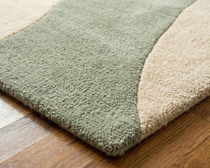 May include: Close-up of a soft, textured rug with a wavy design. The rug features a light green section and a beige section, creating a contrasting pattern. The rug is placed on a wooden floor.