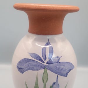 May include: A ceramic vase with a white body and a terracotta-colored rim. The vase is decorated with a blue iris flower and green leaves. The vase is approximately 6 inches tall.