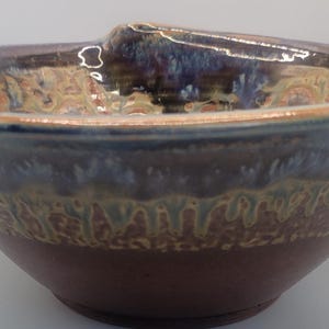 May include: A handcrafted ceramic bowl with a unique, organic shape. The bowl features a rich color palette of deep brown, blue, and tan, with a textured, patterned band around the middle. The rim has small handles. This decorative bowl is ideal for serving or display.