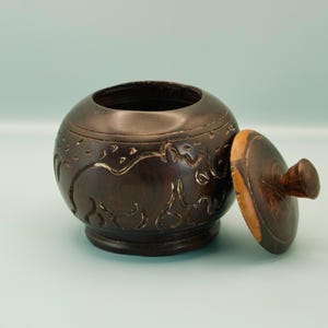 May include: A dark brown, round wooden bowl with a lid. The bowl has carved designs around the sides and a matching lid with a small knob. The lid is partially open, revealing the bowl's interior. The bowl sits on a small base.