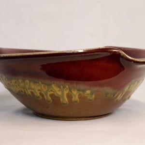 May include: A handmade ceramic bowl with a glossy, deep red interior and a textured, earthy brown exterior. The rim has a unique, wavy design. The bowl's exterior features a band of green and yellow accents. This decorative bowl is perfect for serving.