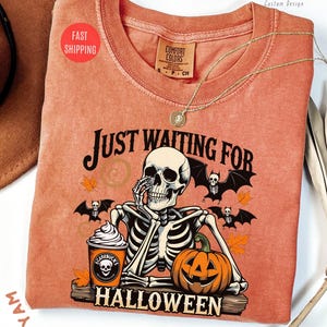 May include: Orange Comfort Colors t-shirt with a Halloween-themed graphic. The design features a skeleton, bats, a pumpkin, and the text "JUST WAITING FOR HALLOWEEN." A coffee cup with a skull logo is also visible.