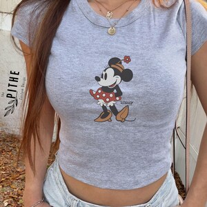 May include: A grey crop top with a Minnie Mouse graphic. The cartoon character is wearing a red polka dot skirt and a flower in her hair. The text "Disney" is below the graphic. The shirt is labelled "ATHLETIC HEATHER".