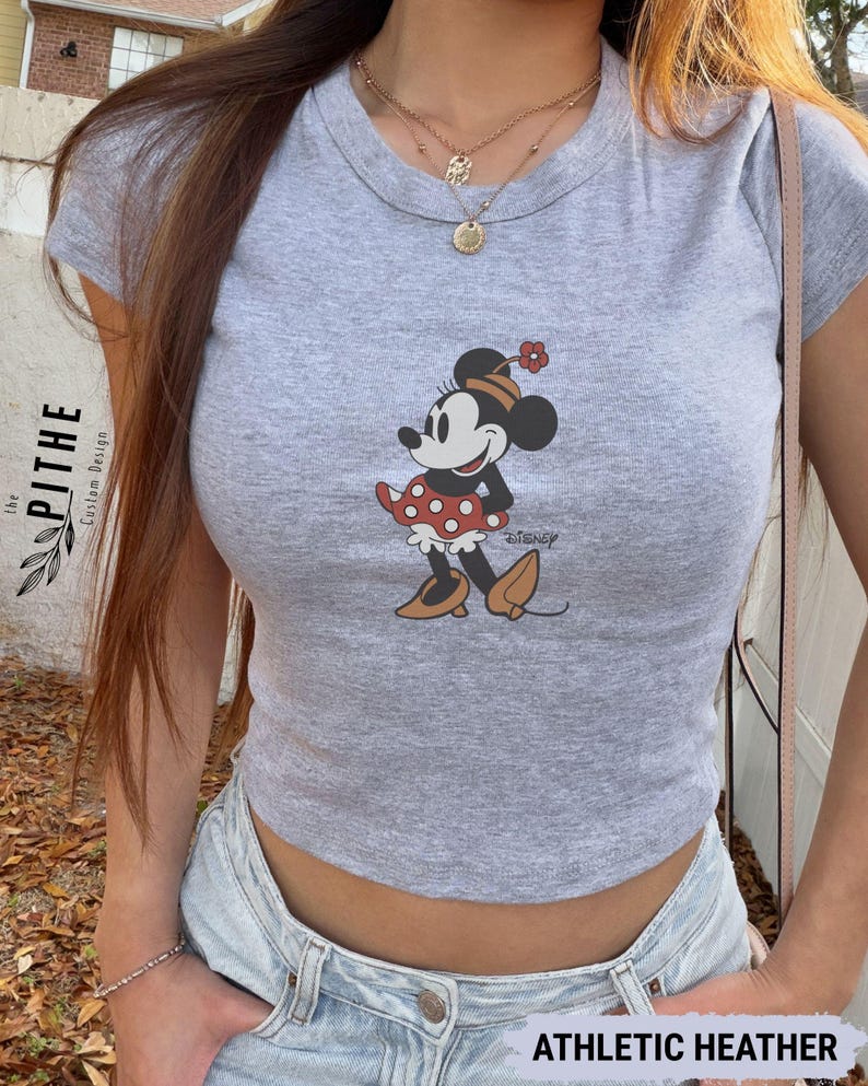 May include: A grey crop top with a Minnie Mouse graphic. The cartoon character is wearing a red polka dot skirt and a flower in her hair. The text "Disney" is below the graphic. The shirt is labelled "ATHLETIC HEATHER".