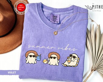 May include: Violet Comfort Colors t-shirt with a 'summer vibes' graphic. The design features three cartoon ghosts wearing sunglasses and hats, enjoying summer activities. A gold necklace and a brown hat are also visible.
