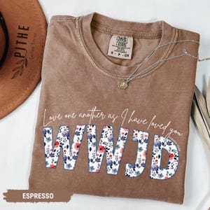 K&ouml;nnte beinhalten: Espressofarbenes T-Shirt mit dem Text "Love one another as I have loved you." und "WWJD" in floralem Design. Eine silberne Halskette mit Anh&auml;nger liegt auf dem Shirt. Das Shirt hat ein Comfort Colors-Etikett.