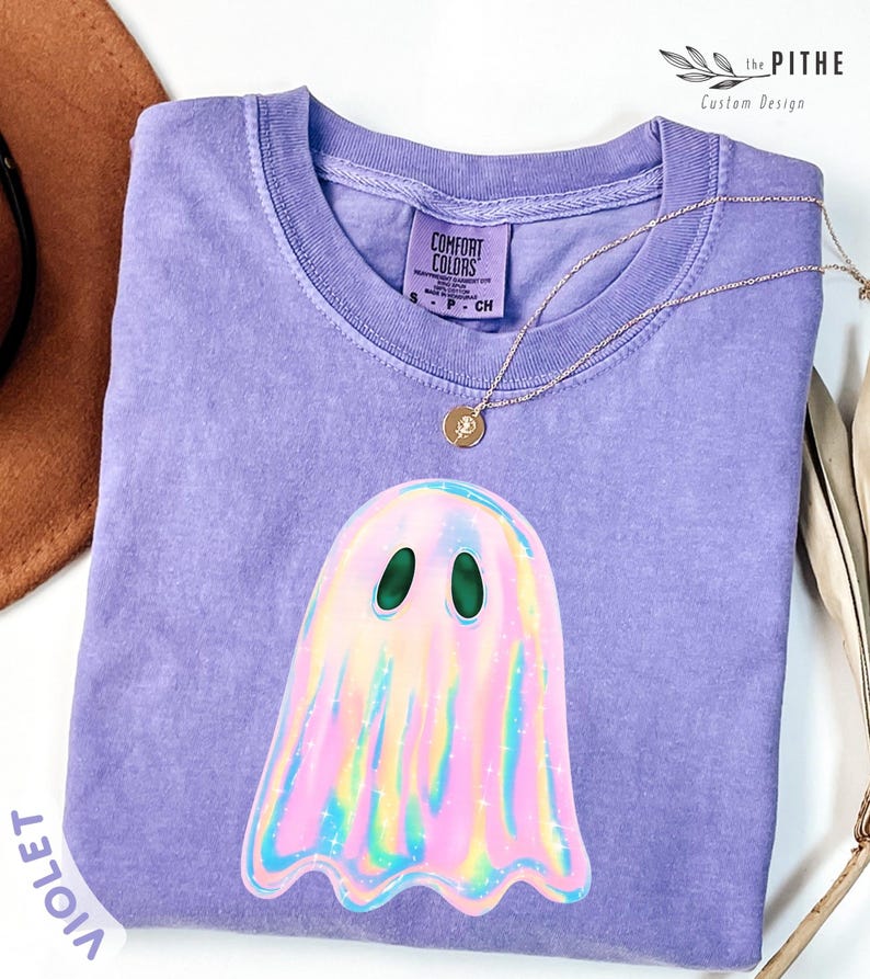 May include: A violet Comfort Colors t-shirt featuring a holographic ghost graphic. The ghost has green eyes and a colorful, iridescent design. A gold necklace with a small pendant rests on the shirt. The shirt is folded and displayed.