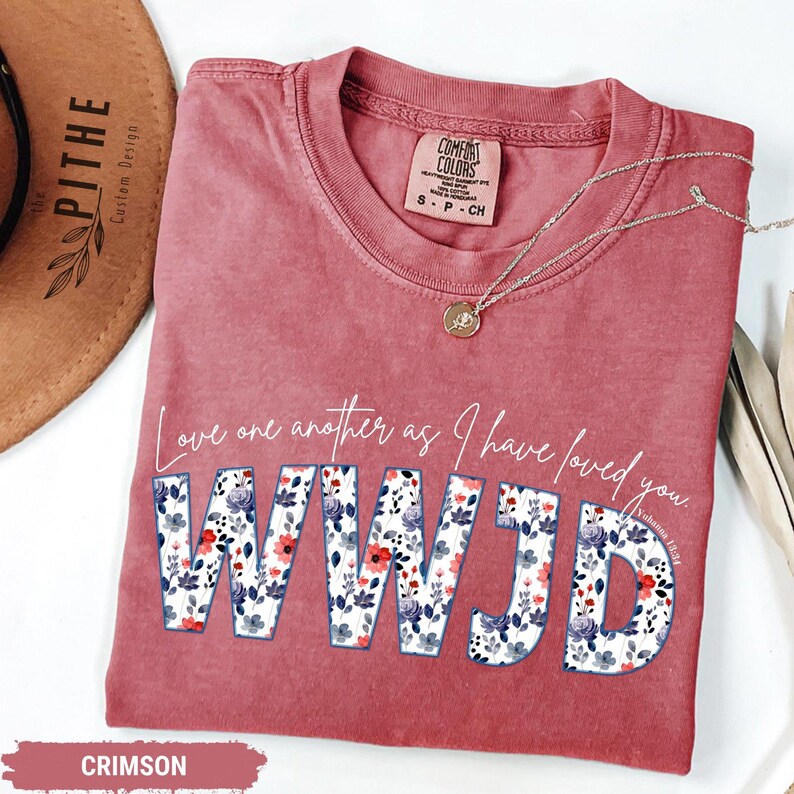 K&ouml;nnte beinhalten: Karmesinrotes T-Shirt mit dem Text "Love one another as I have loved you. WWJD" in Wei&szlig; und floralem Druck. Eine goldene Halskette mit Anh&auml;nger liegt auf dem Shirt. Das Shirt ist von der Marke Comfort Colors, Gr&ouml;&szlig;e S-CH.