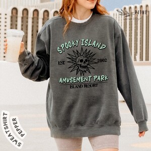 May include: A dark gray sweatshirt with the text "Spooky Island Amusement Park Island Resort" and a skull graphic. The text is in a light green color. The sweatshirt has long sleeves and a crew neck.