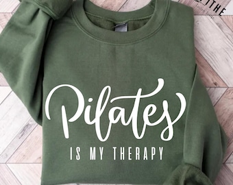 May include: Military green sweatshirt with the white text "Pilates is my therapy." The long-sleeved top has a crew neck and is made of a soft material. The text is in a cursive font. The sweatshirt is a casual style.