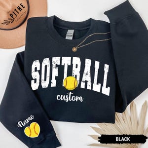 Może przedstawiać: Czarna bluza z napisem "SOFTBALL" w białych, zniszczonych literach, żółtą grafiką do softballu i słowem "custom" poniżej. Lewy rękaw ma grafikę do softballu i słowo "Name".