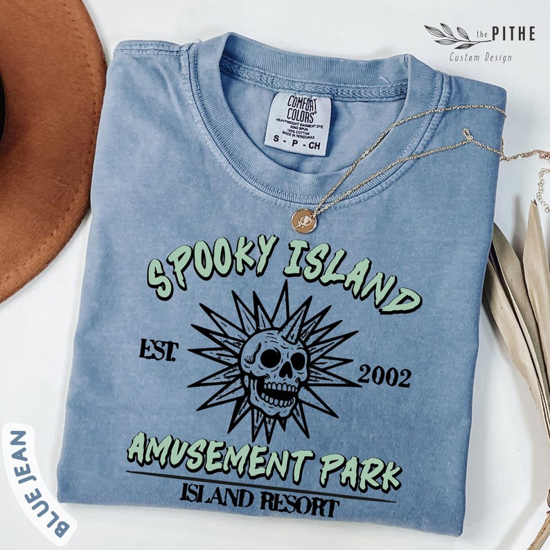 May include: Blue jean colored t-shirt with the text "SPOOKY ISLAND AMUSEMENT PARK ISLAND RESORT" and a skull graphic. The shirt has a crew neck and short sleeves. A gold necklace is draped across the shirt.