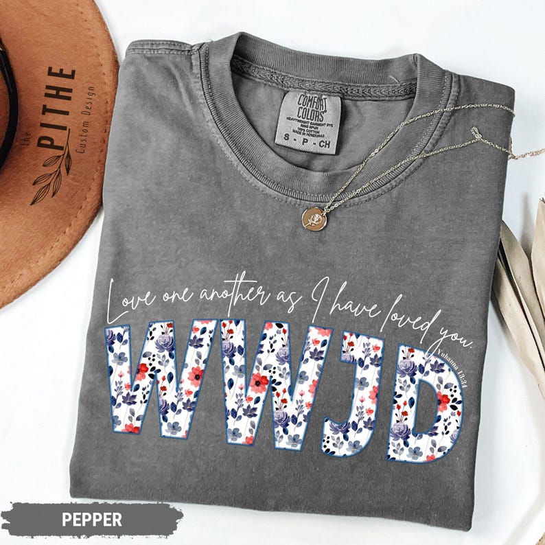 K&ouml;nnte beinhalten: Ein graues Comfort Colors T-Shirt mit dem Text "Love one another as I have loved you. WWJD" in wei&szlig;er Schrift und floralen Buchstaben. Eine silberne Halskette mit Anh&auml;nger liegt auf dem Shirt. Das Shirt ist mit "Pepper" gekennzeichnet.