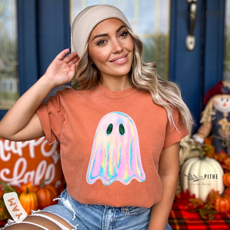 May include: An orange short-sleeved t-shirt featuring a holographic ghost design with green eyes. The shirt is part of a Halloween-themed collection. The model is wearing a beige beanie.