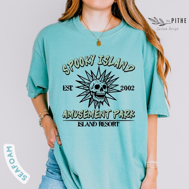 May include: A seafoam green t-shirt with the text "Spooky Island Amusement Park Island Resort" in a black and light green design. The design includes a skull and sunburst graphic. The shirt has short sleeves.