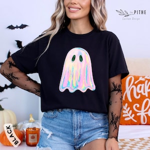 May include: Black t-shirt featuring a holographic ghost graphic with green eyes. The ghost design has a shimmering, iridescent effect. The shirt is part of a Halloween-themed collection.