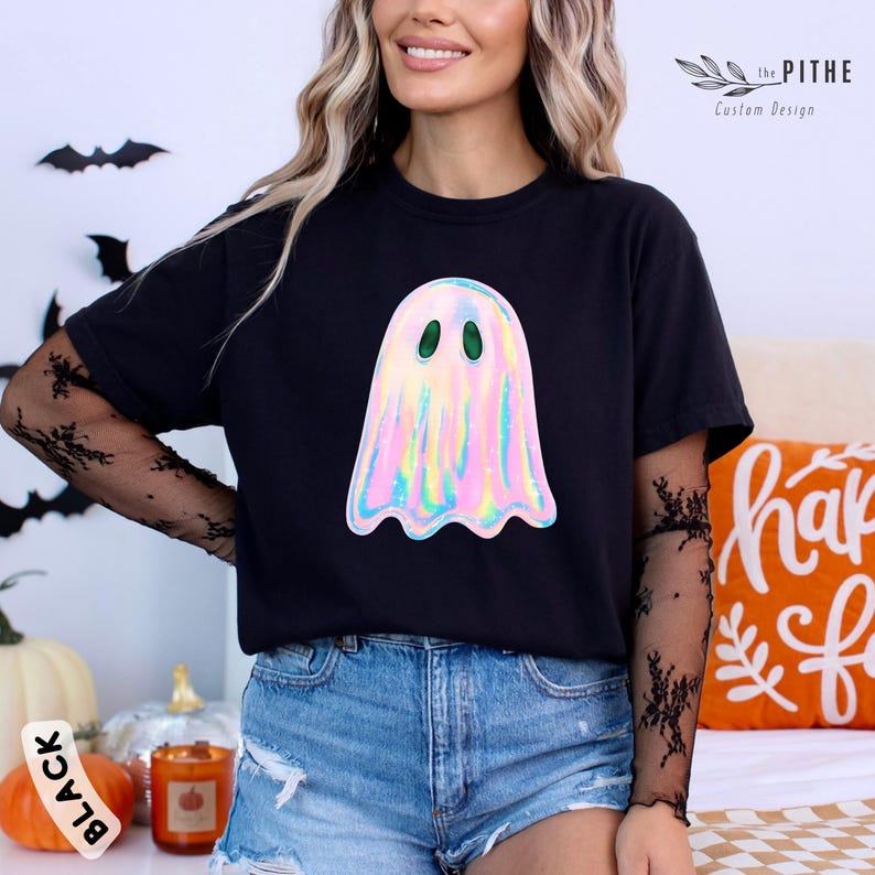 May include: Black t-shirt featuring a holographic ghost graphic with green eyes. The ghost design has a shimmering, iridescent effect. The shirt is part of a Halloween-themed collection.