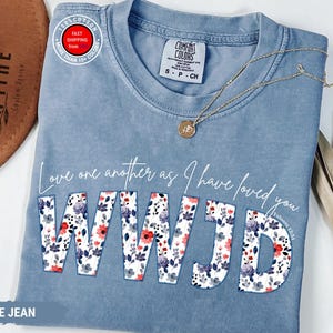 K&ouml;nnte beinhalten: Blaues Jeans-T-Shirt mit dem Text "Love one another as I have loved you. WWJD" in wei&szlig;er Schrift und floralem Druck. Eine goldene Halskette mit Anh&auml;nger liegt auf dem Shirt. Ein brauner Hut ist im Hintergrund zu sehen.