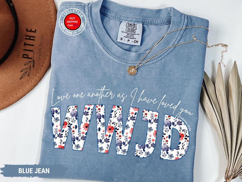 K&ouml;nnte beinhalten: Blaues Jeans-T-Shirt mit dem Text "Love one another as I have loved you. WWJD" in wei&szlig;er Schrift und floralem Druck. Eine goldene Halskette mit Anh&auml;nger liegt auf dem Shirt. Ein brauner Hut ist im Hintergrund zu sehen.