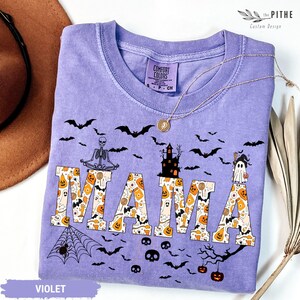 以下が含まれることがあります： ハロウィーンをテーマにしたデザインの、紫色のComfort Colors Tシャツ。「MAMA」の文字はカボチャ、お化け、キャンディーコーンで埋められています。コウモリ、骸骨、お化け屋敷も特徴です。金のネックレスがシャツの上にあります。