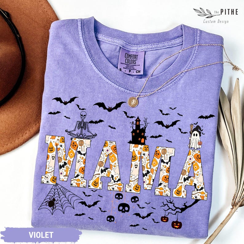 以下が含まれることがあります： ハロウィーンをテーマにしたデザインの、紫色のComfort Colors Tシャツ。「MAMA」の文字はカボチャ、お化け、キャンディーコーンで埋められています。コウモリ、骸骨、お化け屋敷も特徴です。金のネックレスがシャツの上にあります。