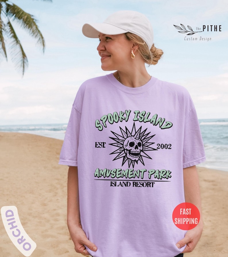 May include: A light purple t-shirt with the text "SPOOKY ISLAND AMUSEMENT PARK ISLAND RESORT" and a skull graphic. The shirt has a relaxed fit and is being worn by a person on a beach. The shirt also has the text "EST 2002".