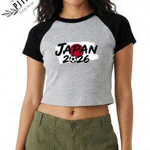 Może przedstawiać: Szara i czarna koszulka typu crop top w stylu baseballowym z napisem "JAPAN" w czarnym kolorze, czerwonym okręgiem i numerem "2026" z grafiką piłki nożnej. Koszulka ma czarne rękawy i czarny kołnierz.