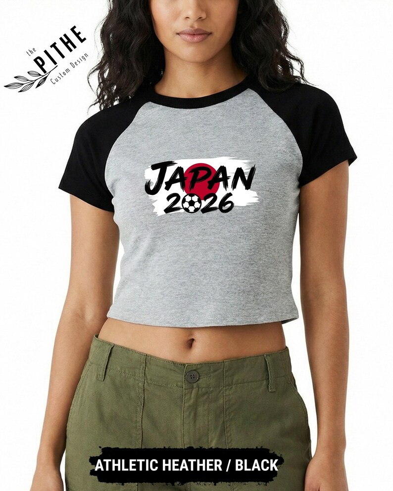 Może przedstawiać: Szara i czarna koszulka typu crop top w stylu baseballowym z napisem "JAPAN" w czarnym kolorze, czerwonym okręgiem i numerem "2026" z grafiką piłki nożnej. Koszulka ma czarne rękawy i czarny kołnierz.