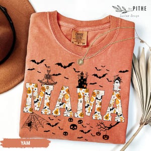 以下が含まれることがあります： ハロウィーンをテーマにしたオレンジ色のTシャツ。装飾的なフォントで「MAMA」の文字がハロウィーンのアイコンで埋められています。シャツには、コウモリ、スケルトン、お化け屋敷、幽霊が描かれています。金のネックレスがシャツの上にかけられています。