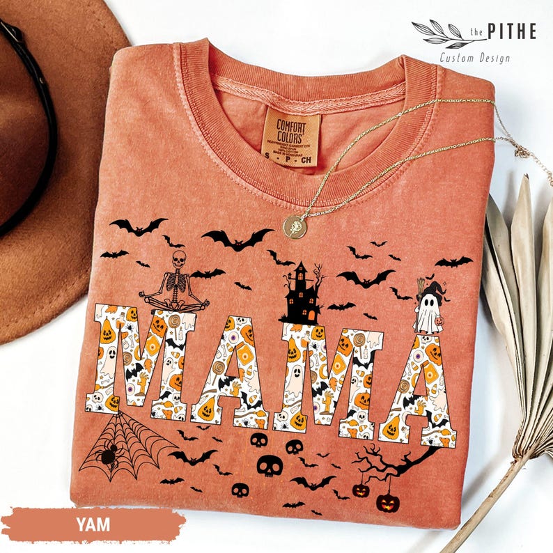 以下が含まれることがあります： ハロウィーンをテーマにしたオレンジ色のTシャツ。装飾的なフォントで「MAMA」の文字がハロウィーンのアイコンで埋められています。シャツには、コウモリ、スケルトン、お化け屋敷、幽霊が描かれています。金のネックレスがシャツの上にかけられています。