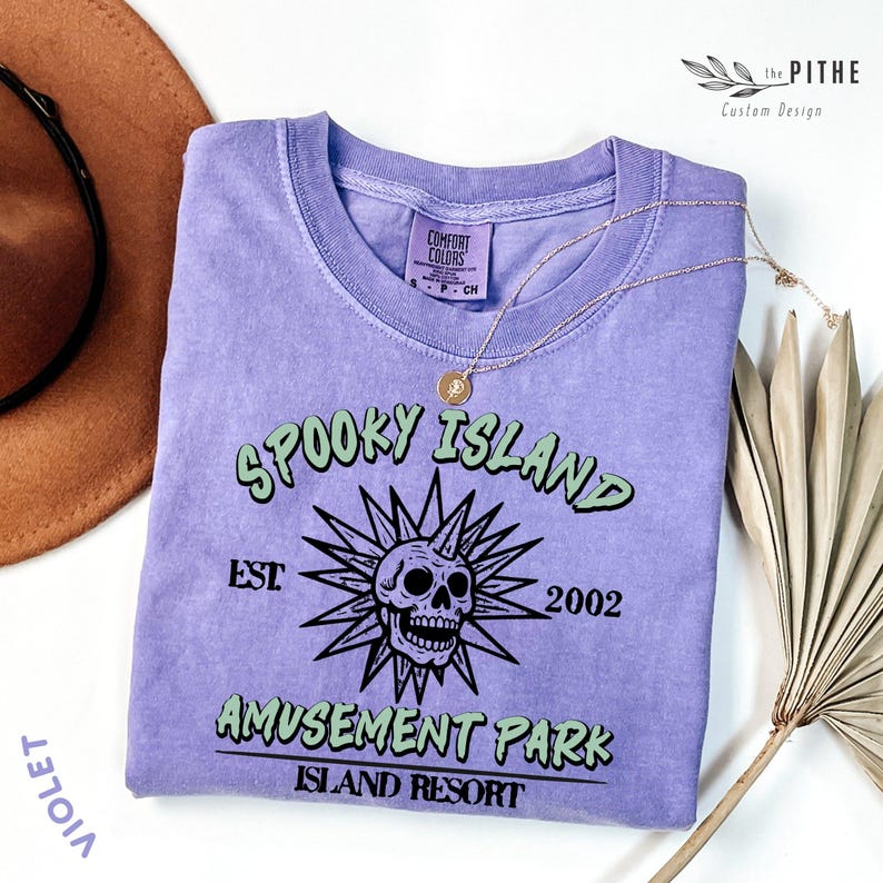 May include: A violet-colored t-shirt with the text "SPOOKY ISLAND AMUSEMENT PARK ISLAND RESORT" and a skull graphic. The shirt has a crew neck and short sleeves. A gold necklace with a pendant rests on the shirt.