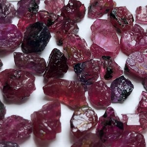 Puede incluir: Una colección de gemas en bruto, sin cortar, en tonos de rojo intenso, púrpura y rosa. Las piedras varían en tamaño y forma, con una textura rugosa y natural. Las gemas están dispersas sobre una superficie blanca.