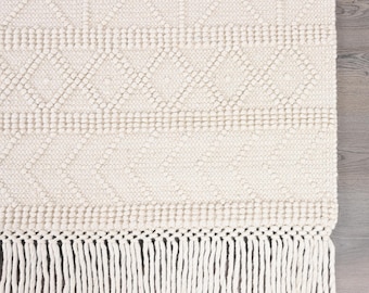 May include: An off-white woven rug featuring a textured geometric pattern of diamonds and horizontal bands. The rug is finished with a long, knotted fringe along the bottom edge, creating a tactile surface with a consistent cream-colored yarn.