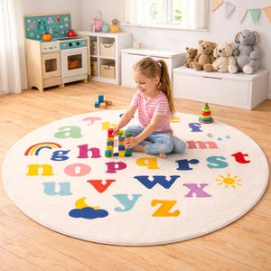 May include: A round, off-white children's rug featuring the alphabet in various bright colors. A rainbow, sun, and cloud are also depicted. A young child is playing with colorful blocks on the rug in a playroom setting.