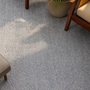May include: A blue and white patterned rug with a herringbone design. The rug has a light blue and white colour scheme and is made of a soft, textured material. The rug is in a room with wooden furniture and a potted plant.