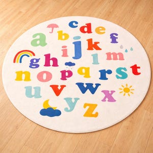May include: A round, off-white rug featuring the alphabet in various bright colors. Each letter is distinct, with accompanying illustrations like a rainbow, sun, and cloud. The rug is designed for a child's room or play area.
