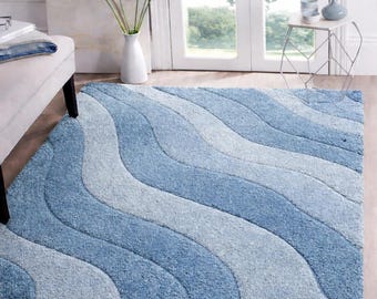 May include: A rectangular area rug featuring a wavy, multi-tonal pattern of light blue, medium blue, and soft grey. The rug has a thick, textured pile with a consistent design of flowing, curved stripes that span across the entire surface area.