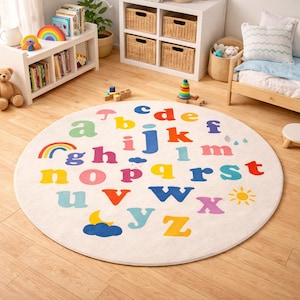 May include: A round, off-white children's rug featuring the alphabet in various bright colors. The letters are scattered across the rug, with illustrations of a rainbow, sun, moon, and clouds. The rug is placed on a light wood floor.