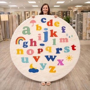 May include: A round, off-white rug featuring the alphabet in various bright colors. The rug includes illustrations of a rainbow, sun, moon, and rain. The rug is held by a person in a store setting.