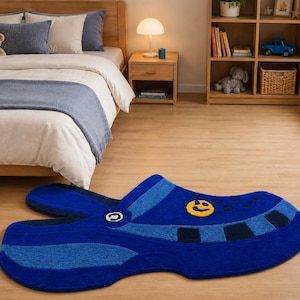 May include: A blue rug shaped like a clog shoe with a textured surface. The rug features a bright yellow smiley face icon on the side, dark blue rectangular accents along the edge, and a small white circular detail near the heel area.