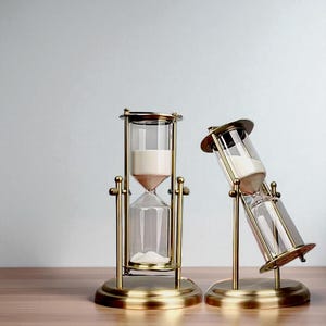 以下が含まれることがあります： 真鍮製の砂時計が2つ。砂時計は透明なガラスボディと真鍮フレームで構成されています。1つの砂時計は直立しており、もう1つは傾いています。砂は白とピンク色です。
