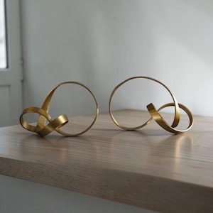 Puede incluir: Dos esculturas abstractas de color dorado. Cada escultura presenta una forma circular con un elemento en forma de cinta que se curva y se entrelaza. Las esculturas se exhiben sobre una superficie de madera clara, sobre un fondo neutro.